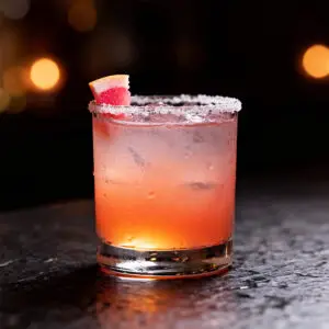 Angled photo of a grapefruit margarita with a grapefruit wedge and salted rim, moody dark bar background.