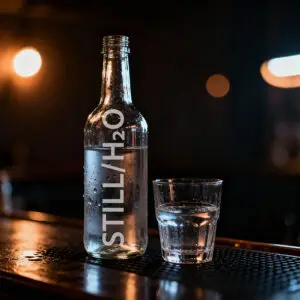 Unlabelled glass bottle of still water beside a matching glass half-filled with water, condensation on both, dark bar background with warm bokeh lighting.